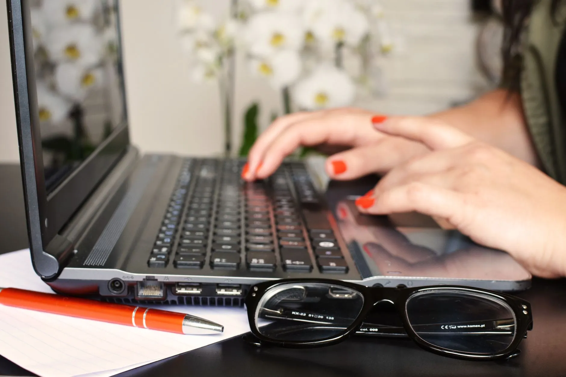 Mãos digitando em um notebook ao lado de um caderno e de óculos, escrevendo a carta de apresentação