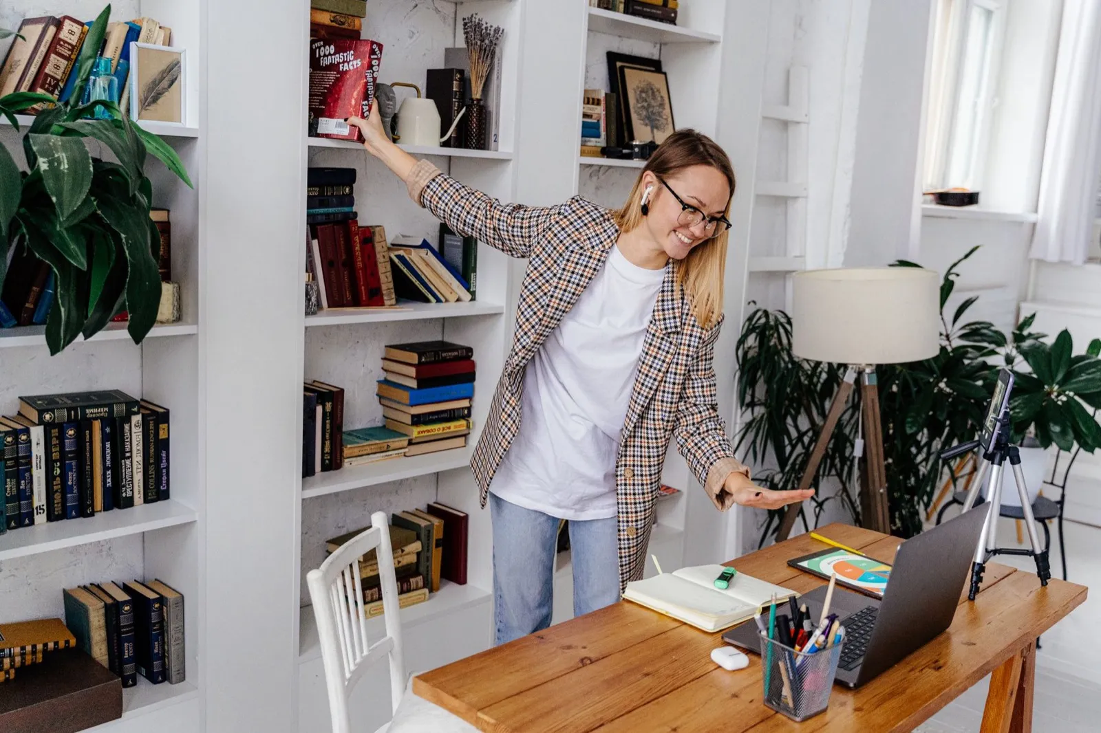 Mulher pegando um livro na estante ao lado da mesa com notebook no escritório de casa